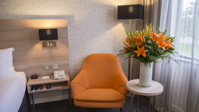 Edge of bed on left with orange comfortable sofa chair next to window with flowers on table