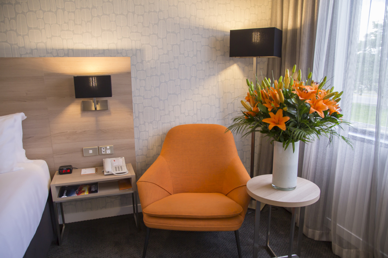 Edge of bed on left with orange comfortable sofa chair next to window with flowers on table