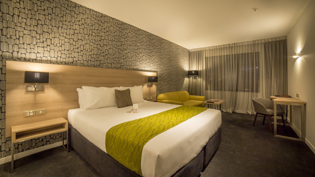 Luxury room with large bed, side table, sofa, table, flatscreen TV, desk and chair with lamp in corner.