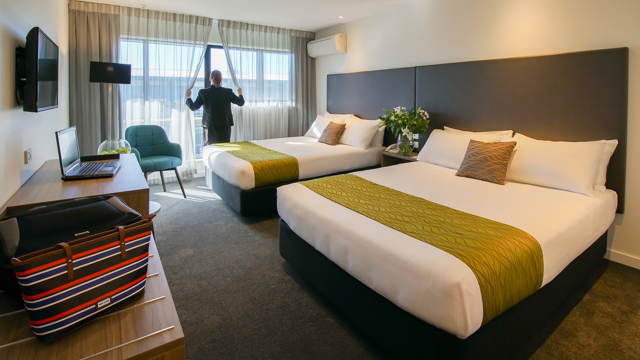 Two large beds with lounge chair, desk and table, TV and lamp in corner.