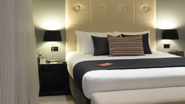 Large bed with luxury linen, headboard, lamps and ottoman seat.