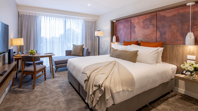 Luxury room with quality bed and furnishings, art on the wall, table and chairs, TV with chaise lounge next to large windows.