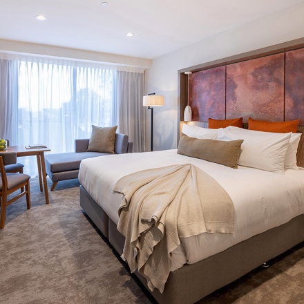 Luxury room with quality bed and furnishings, art on the wall, table and chairs, TV with chaise lounge next to large windows.