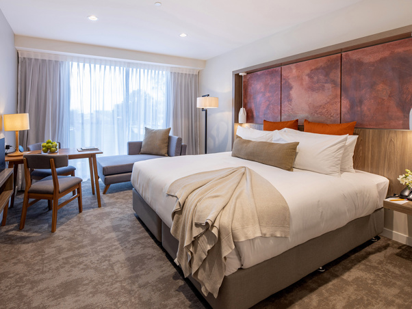 Luxury room with quality bed and furnishings, art on the wall, table and chairs, TV with chaise lounge next to large windows.