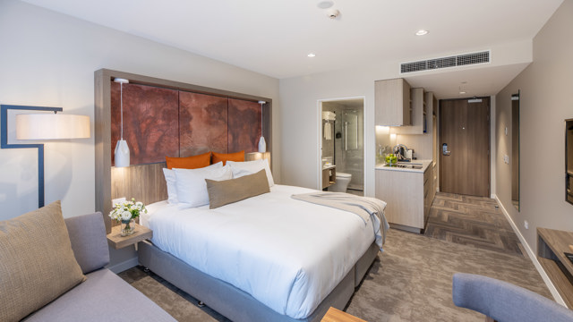 Luxury room showing bed and furnishings, art on the wall, kitchen area, table and chairs, TV with chaise lounge next to large windows.