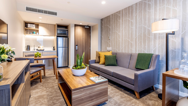 Lounge space with kitchenette area. Sofa, dining table and chairs, coffee table in centre, flatscreen on wall.