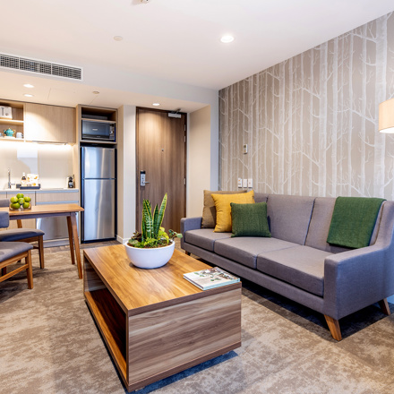 Lounge space with kitchenette area. Sofa, dining table and chairs, coffee table in centre, flatscreen on wall.
